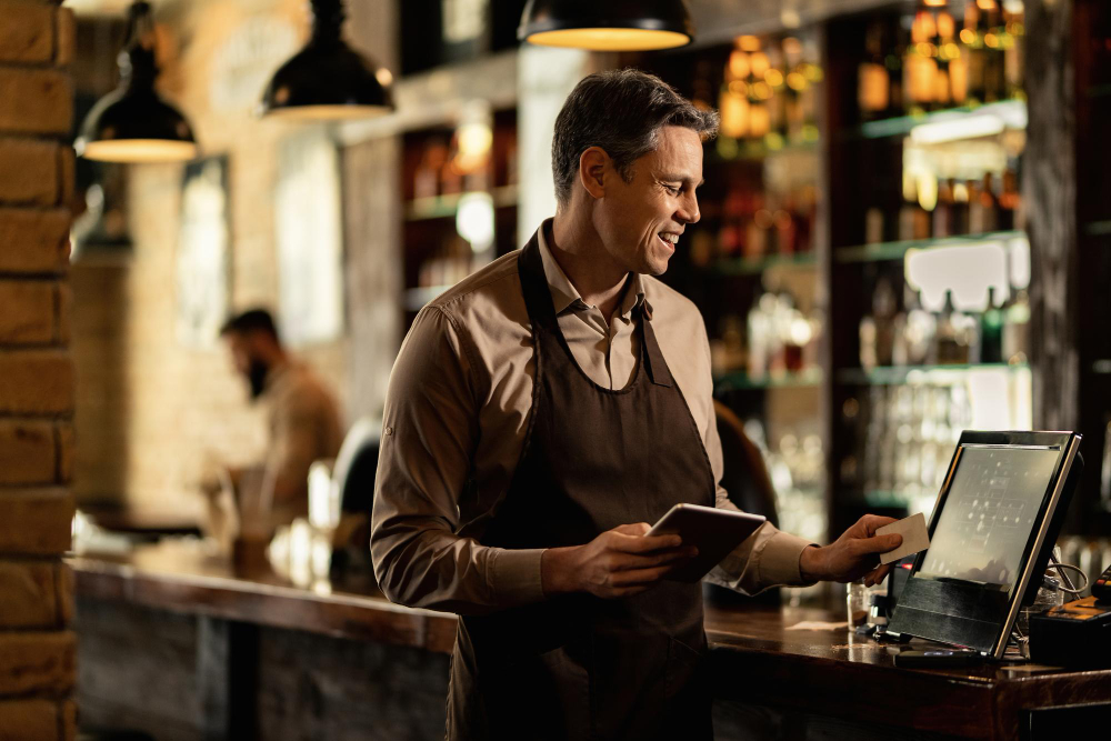 Smiling restaurant manager holding a tablet while processing a payment at the bar counter