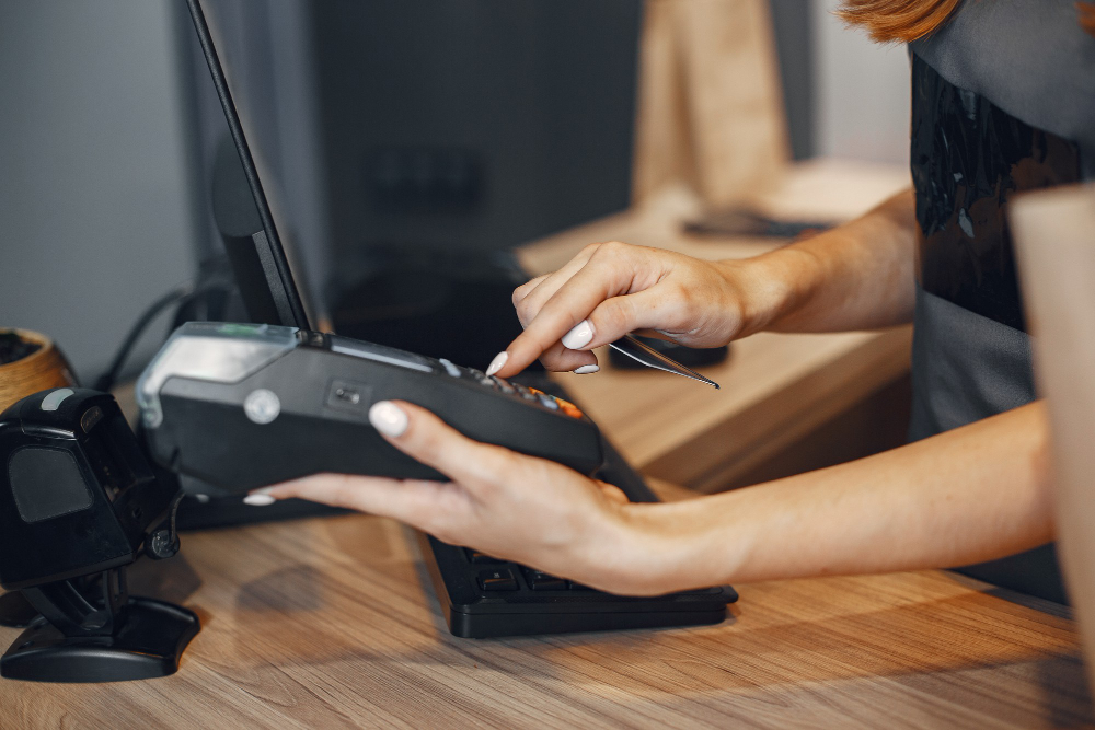 Cashier holding a card payment terminal at a wooden restaurant counter beside a barcode scanner