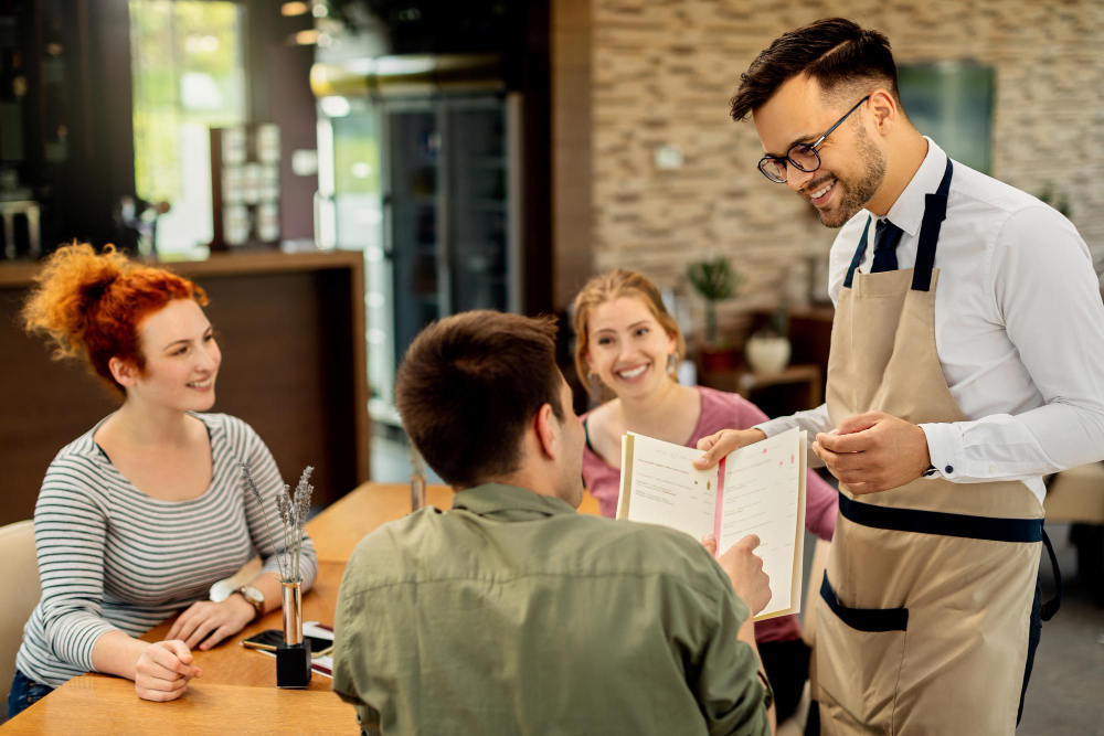 Smiling waiter presenting the menu to a group of friends at a cafe table